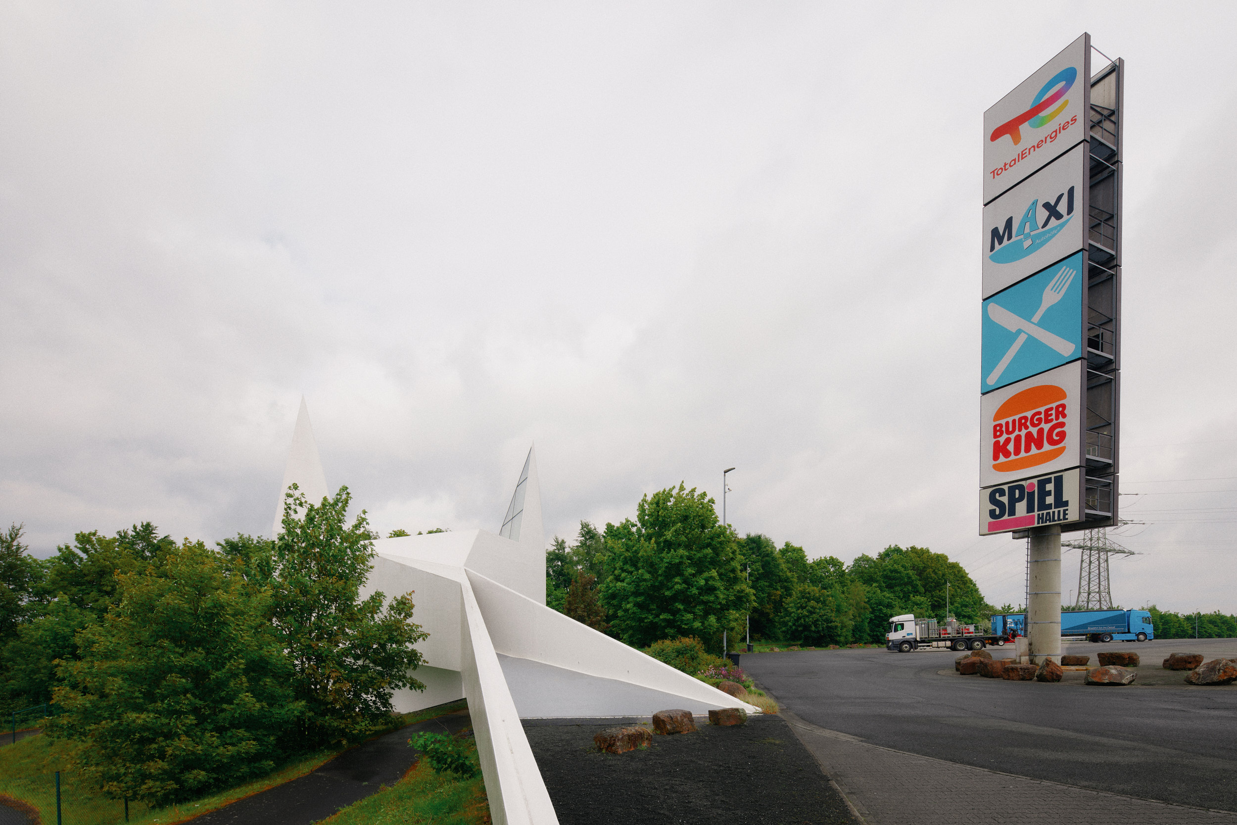 Tall roadside sign with TotalEnergies, Maxi, Burger King, and Spielhalle logos beside a modern white building