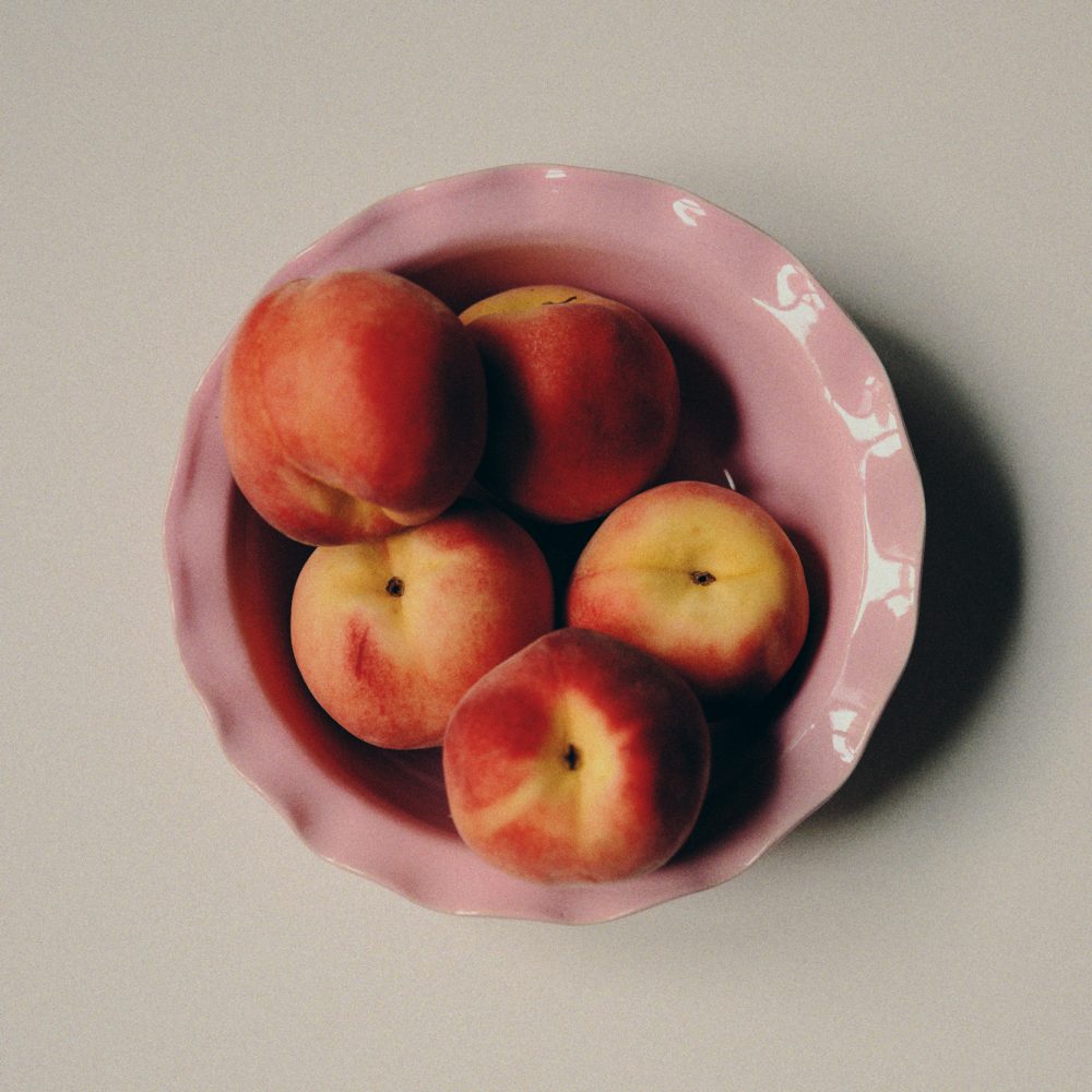 A still life photograph with pink ceramic bowl by Viola Beuscher filled with five peaches on a light background as a fine art print