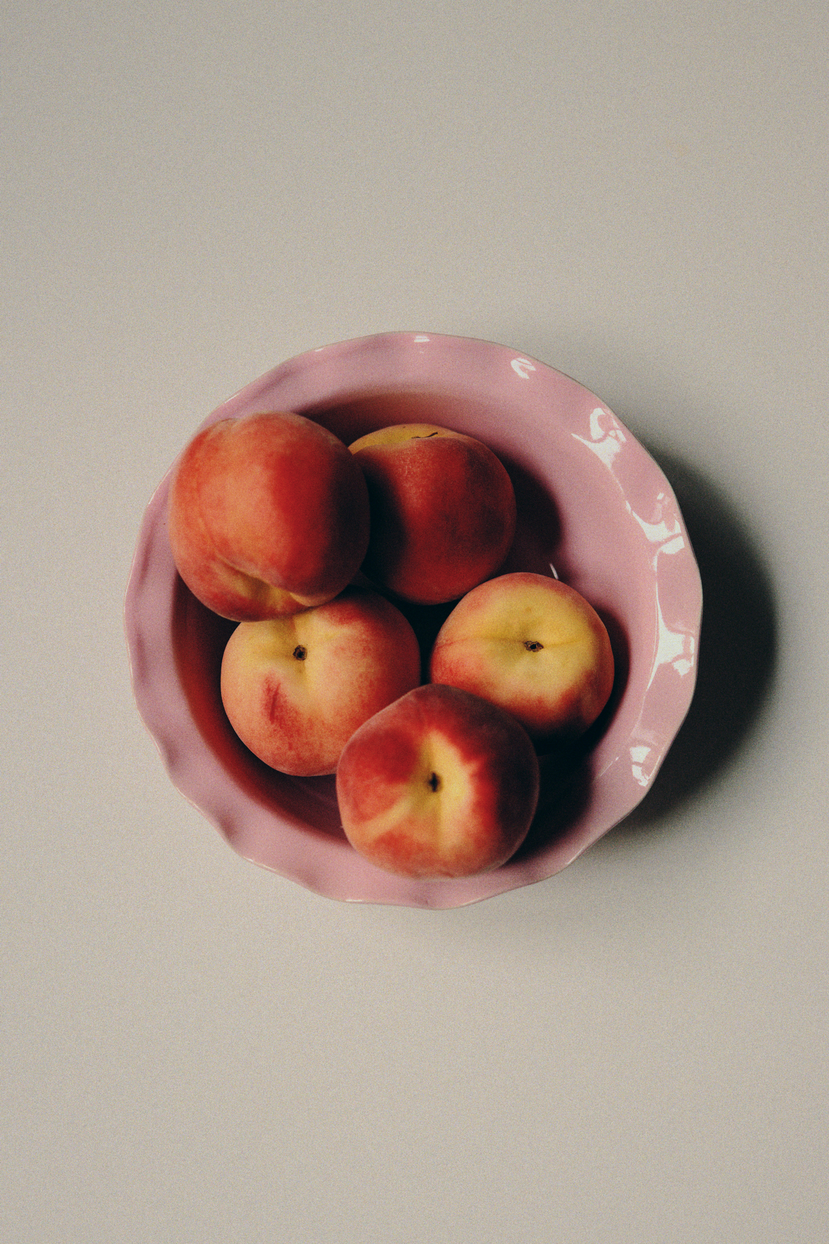 A still life photograph with pink ceramic bowl by Viola Beuscher filled with five peaches on a light background as a fine art print